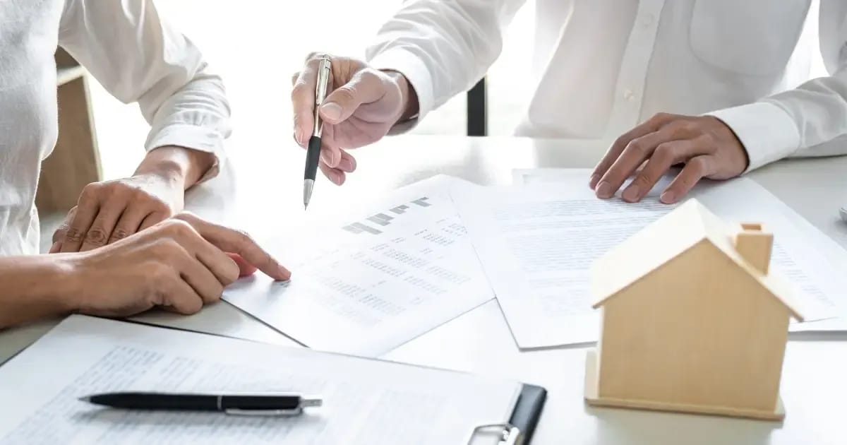 Two people discussing and highlighting a roof insurance claim document with a wooden model of a house sitting on the desk between them. 